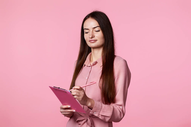 beautiful brunette business woman holding clipboard with papers document on pink background. - Foto, Bild