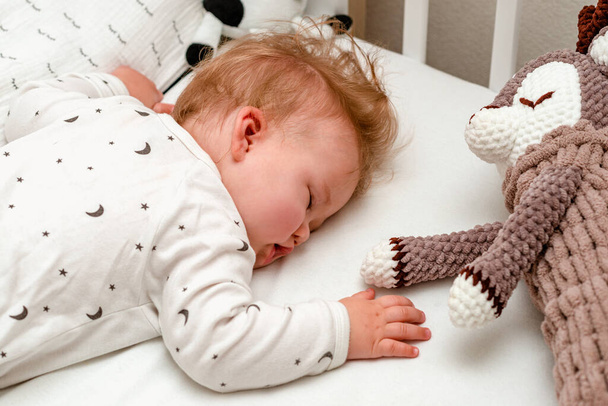 Nahaufnahme Porträt eines blonden Babys, das ruhig schläft, mit Stofftier auf dem Bauch in einem Kinderbett. - Foto, Bild