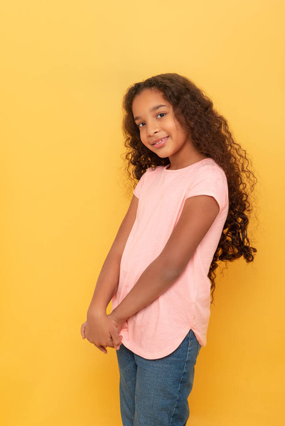 Портрет темношкірої молодої усміхненої дівчини з довгим кучерявим волоссям у рожевій сорочці на жовтому тлі
 - Фото, зображення