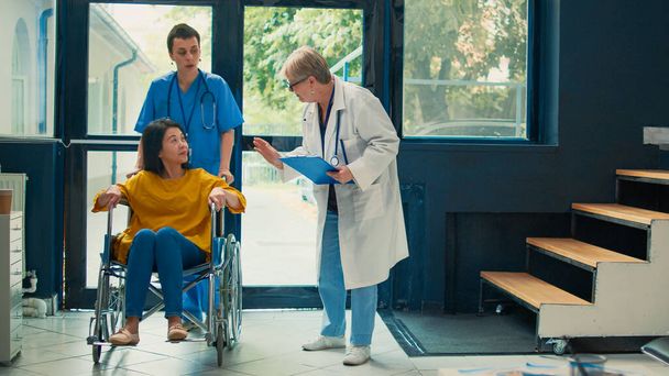 Asystent medyczny pomaga pacjentowi na wózku inwalidzkim z zaburzeniami, uczestniczy w wizycie w ośrodku zdrowia. Kobieta na wózku inwalidzkim cierpiąca na przewlekłą niepełnosprawność fizyczną w lobby. - Zdjęcie, obraz