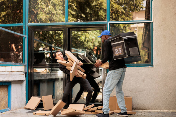 Verwarde food delivery service klant vangen vallende pizza dozen, roekeloze koerier droporder in de buurt van kantoorgebouw buiten. Onvoorzichtige man leveren fastfood pakketten stapel - Foto, afbeelding