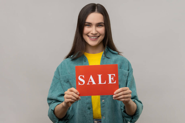 Portrait of young adult happy smiling woman standing holding card with sale inscription, huge discounts, wearing casual style jacket. Indoor studio shot isolated on gray background. - Fotoğraf, Görsel