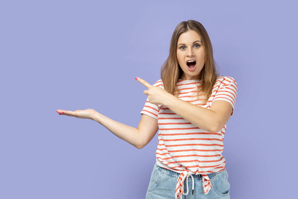 Portrait of surprised astonished blond woman wearing striped T-shirt presenting advertising area on her palm and pointing to copy space. Indoor studio shot isolated on purple background - Photo, image