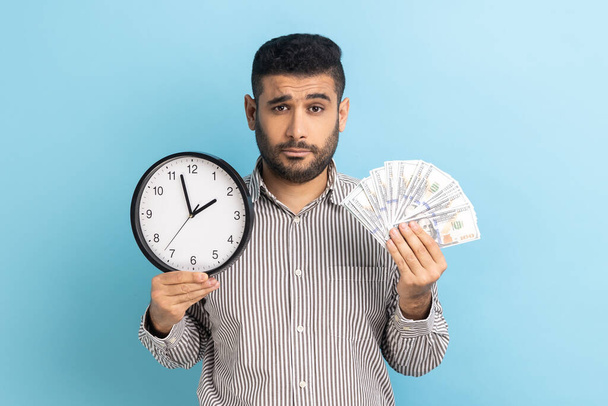 Portrait of upset sad businessman holding big fan of dollar banknotes and wall clock, time is money, looking at camera, wearing striped shirt. Indoor studio shot isolated on blue background. - Foto, Imagem