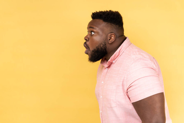 Wow, unbelievable. Side view of man wearing pink shirt has shocked expression, looking at camera with open mouth and amazed big eyes. Indoor studio shot isolated on yellow background. - Фото, изображение