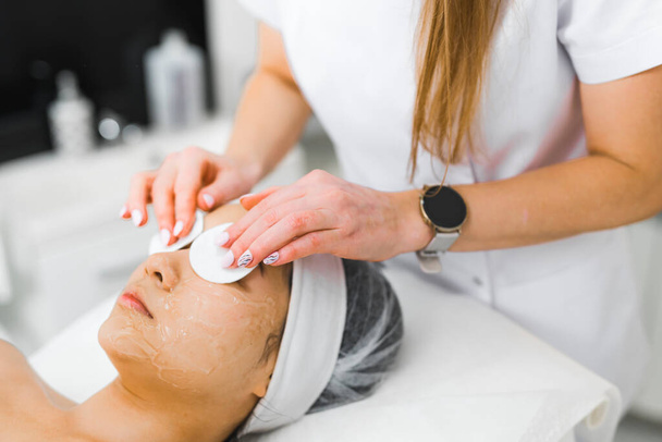 Unrecognizable beautician removing the rest of cleansing mask from the eye area of her young Asian client with the use of cotton cosmetic pads. Skincare concept. . High quality photo - Foto, Bild