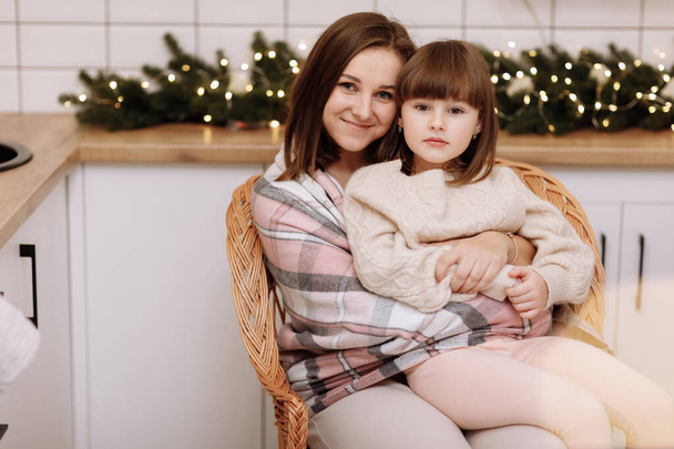 Vrolijk Kerstfeest en Gelukkig Nieuwjaar. Jonge moeder knuffelen en kussen haar dochtertje, zitten en ontspannen samen in rieten stoel in de keuken ingericht voor Kerstmis. Tijd van het familiefeest - Foto, afbeelding