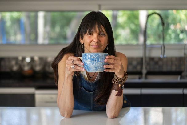 Porträt einer lächelnden Frau, die zu Hause eine entspannte Kaffeepause genießt, während sie in die Kamera blickt. - Foto, Bild