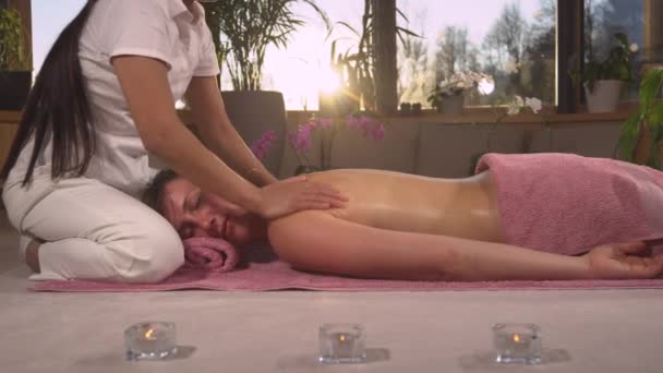 FERMER : Jeune femme relaxante tout en recevant un massage thérapeutique du dos. Les rayons du soleil brillent à travers les mains féminines tout en massant doucement le dos d'une jeune femme. Soin spa en fin de journée - Séquence, vidéo
