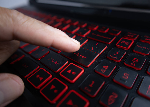 Мужская рука собирается нажать клавишу ввода на клавиатуре ноутбука, с красным освещением. (избирательная направленность субъекта или точки действия намеренно) - Фото, изображение