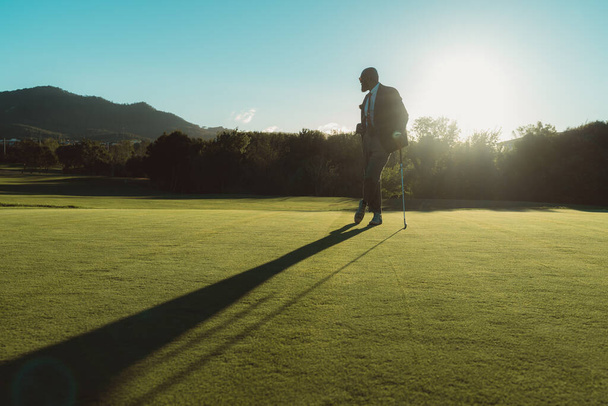 A silhouette of an elegant bald bearded black man in a tailored suit leaning on his gold club while standing on an empty lawn of a golf field, backlit by evening sun casting a long shadow on the grass - Foto, Imagem