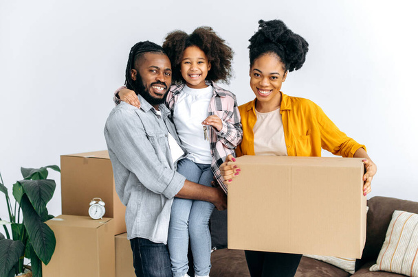 Happy african american family, dad mom and their cute daughter, moved to their new home, holding boxes with things, girl in dads arms shows keys, they smile into camera. New housing, relocation - Photo, Image