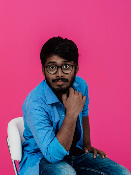 A young Indian student in a blue shirt with glasses sitting in front of a pink background.  - Foto, Bild