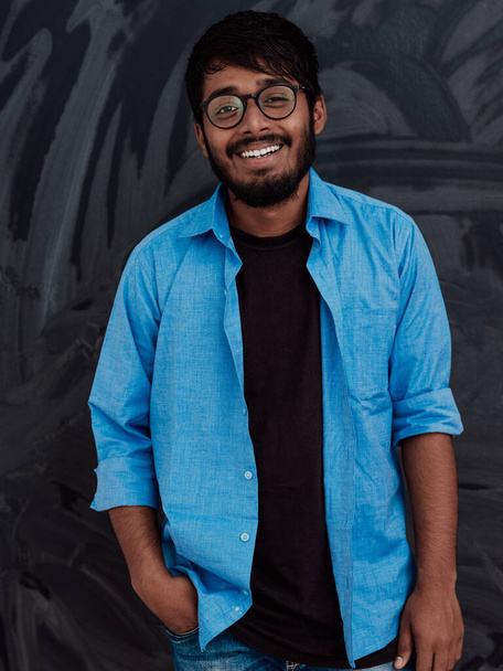 Indian smiling young student in blue shirt and glasses posing on school blackboard background.  - Photo, Image