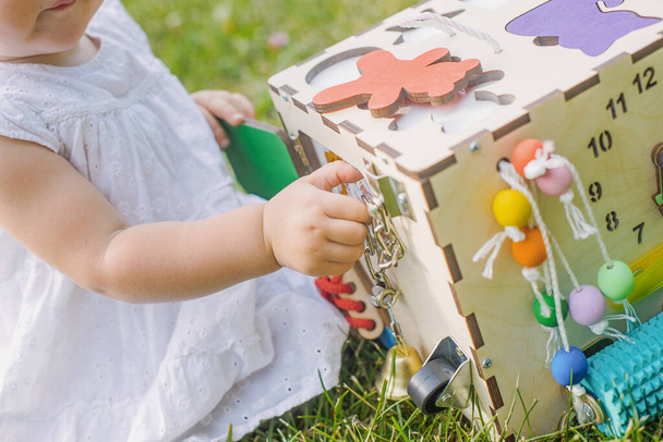 Schattig klein meisje speelt met busiboard buiten op groen gras. Educatief speelgoed voor peuters. meisje opende deur naar kubus van boord - Foto, afbeelding
