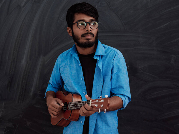Indian young man in a blue shirt and glasses playing the guitar in front of the school blackboard.  - Fotoğraf, Görsel