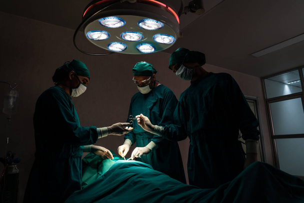 Χειρουργική ομάδα εκτελεί χειρουργική επέμβαση σε ασθενή σε αποστειρωμένο χειρουργείο. Σε ένα χειρουργείο που φωτίζεται από μια λάμπα, μια επαγγελματική και σίγουρη χειρουργική ομάδα παρέχει ιατρική φροντίδα σε έναν αναίσθητο ασθενή.. - Φωτογραφία, εικόνα