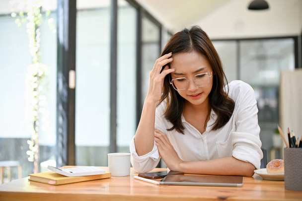 Een gestresste en ernstige duizendjarige Aziatische zakenvrouw of vrouwelijke manager, die lijdt aan depressie en hoofdpijn, zit aan haar bureau. - Foto, afbeelding