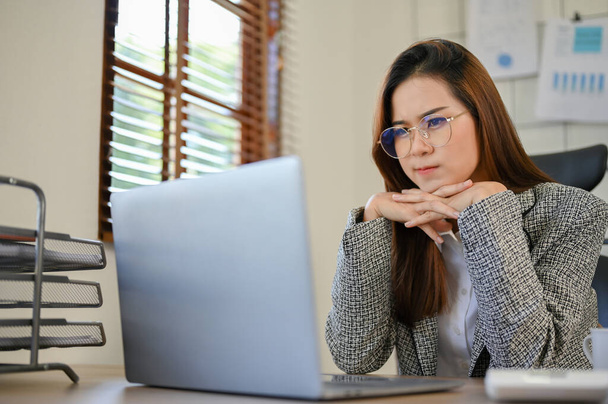 Молода азійська бізнесменка або менеджер жіночої статі в окулярах думає про свій новий проект і шукає натхнення в Інтернеті, використовуючи ноутбук. - Фото, зображення