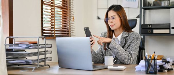 Attraente giovane donna d'affari asiatica o CEO femminile in abito formale aziendale e occhiali seduti sul suo posto di lavoro, utilizzando il suo smartphone per inviare messaggi. - Foto, immagini