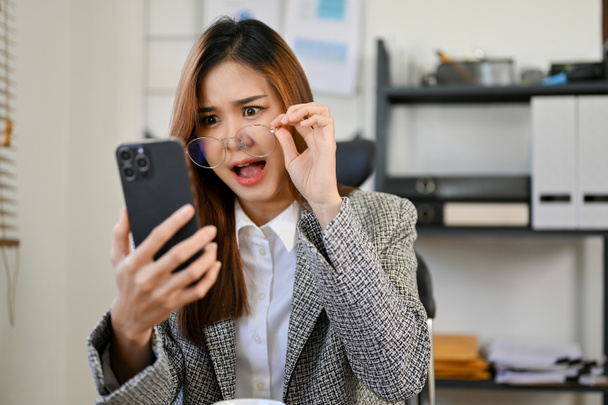 Geschokt en verbaasd jonge Aziatische zakenvrouw aanraken van haar bril, kijken naar haar smartphone scherm, het lezen van een onverwacht nieuws of e-mail.  - Foto, afbeelding