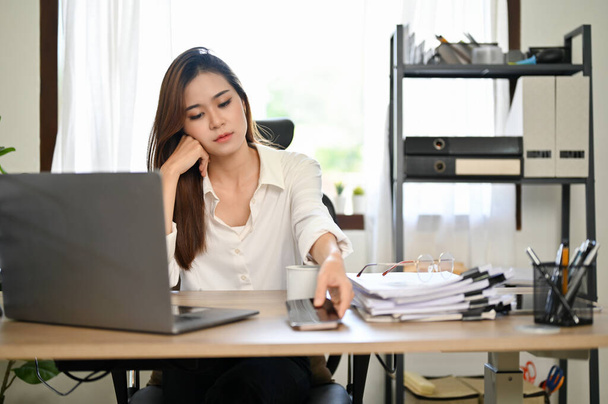Aburrida y cansada empresaria asiática milenaria o gerente se sienta en su escritorio de la oficina, a punto de usar su teléfono inteligente. somnoliento, sin pasión, agotado, con exceso de trabajo. - Foto, Imagen