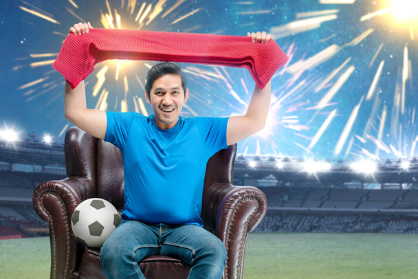 Asian man with the ball holding scarf sitting on the couch with an excited expression in the football stadium background - Foto, afbeelding