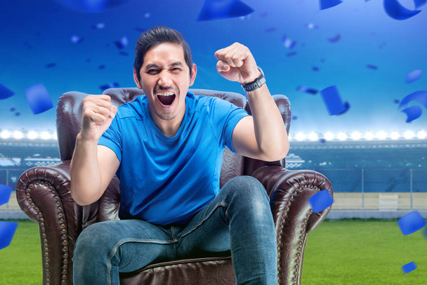 Asian man sitting on the couch with an excited expression in the football stadium background - Foto, immagini