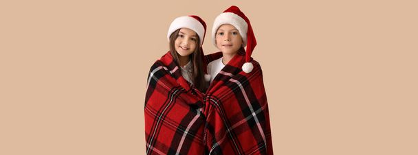 Cute children in pajamas and Santa hats wrapped in plaid on beige background - Fotoğraf, Görsel
