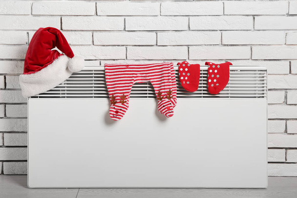 Radiador elétrico com chapéu de Papai Noel, bodysuit bebê e meias perto da parede de tijolo branco - Foto, Imagem