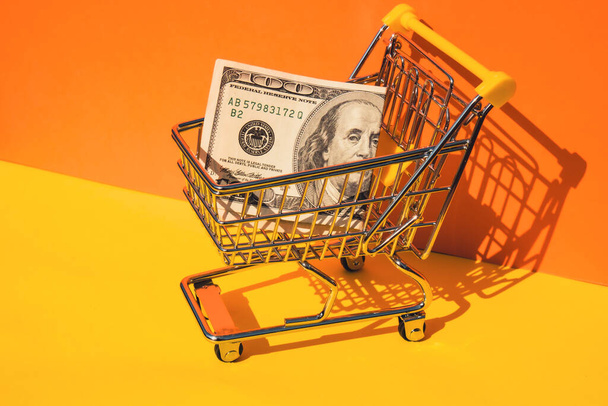 US dollar paper currency 100 banknote money Shopping trolley cart on colorful orange yellow background. Copy space for your text. Online shopping, buy mall market shop consumer concept. Small toy - Photo, Image