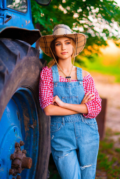 Beautiful blonde cowgirl in straw hat and denim overall standing near blue tractor. Colorful art work. - Foto, Imagen
