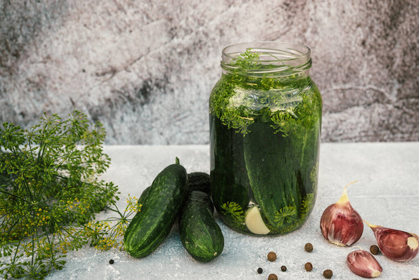 pickling cucumbers for winter organic food, jar with pickled vegetables, garlic, spices, black pepper, dill on gray concrete background - Photo, image