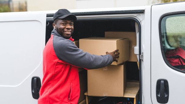 Upcoming holiday season sale via online shopping. Outdoor medium closeup shot of smiling surprised Black man in red-and-gray working clothes taking out one of many cardboard boxes out of his white van - Фото, изображение