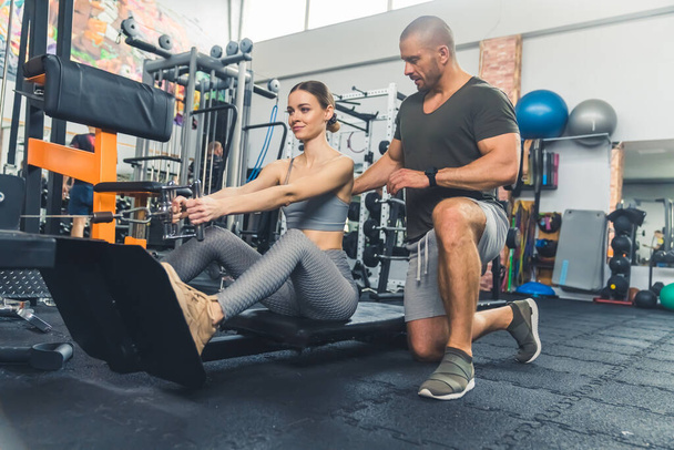 Young, athletic, caucasian woman performing seated back pull on the machine and her man coach helping her to do the exercise correctly in the gym. High quality photo - Photo, Image