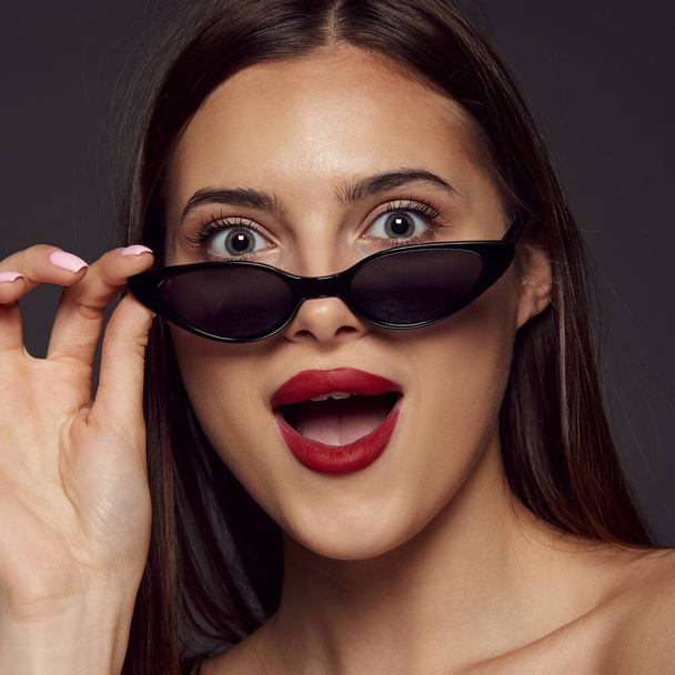 Close-up portrait of young beautiful girl with red lips posing in sunglasses isolated over dark grey background. Emotions. Concept of beauty, skin care, cosmetology, plastic surgery, makeup, fashion - Photo, Image