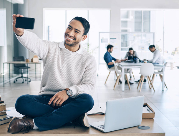 Selfie, phone and businessman sitting on desk in meeting with team, staff and working in corporate office. Startup, happiness and young Asian male worker using smartphone to take picture in workplace. - Foto, afbeelding