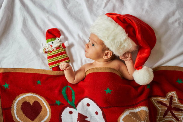 Little newborn baby, wearing Santa hat, lies under red Christmas blanket with toy. Top above view, x-mas home indoors - Fotó, kép