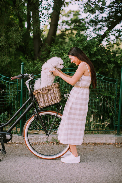 Mujer joven bonita poniendo perro blanco bichon frise en la cesta de la bicicleta eléctrica - Foto, imagen