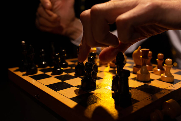 Close-up van het schaakbord met stukken tijdens het toernooi op donkere achtergrond. Selectieve focus op de hand van grootmeester reikend naar zwarte pion. Beeld met dramatische verlichting. Gespannen spel concept - Foto, afbeelding