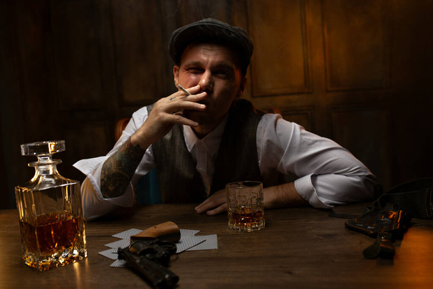 Cinematic portrait of young 1920s British gangster in waistcoat and flat cap sitting at table with cards and gun lying nearby in vintage casino, smoking and drinking whiskey. Peaky blinders style look - Photo, Image