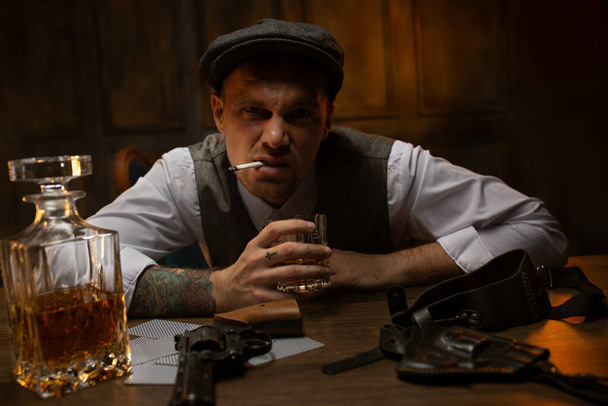 Aggressiivinen nuori gangsteri istuu savuke suussa ja lasi viskiä käsissä vintage kasino, etsivät kurtistaa kameran edessä. Kortit, revolveri ja kotelo makaa edessä mies pöydällä - Valokuva, kuva