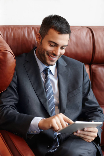My shares are up. A smiling young businessman sitting on a leather sofa while using a digital tablet - Foto, afbeelding