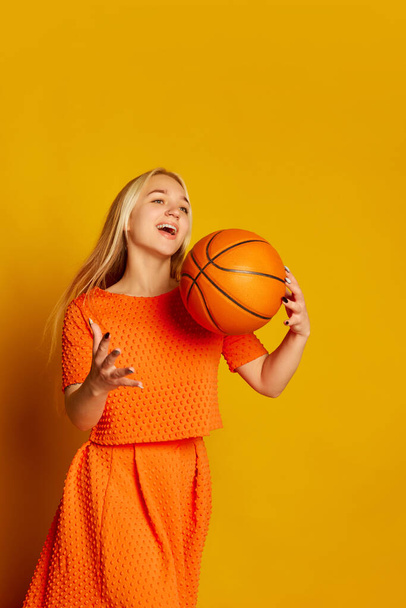 Fun, joy, sport. Young emotional blond girl in orange dress having fun with basketball ball isolated over orange background. Vivid emotions, happiness, hobbies and joy. Looks delighted, excited - Foto, Imagem