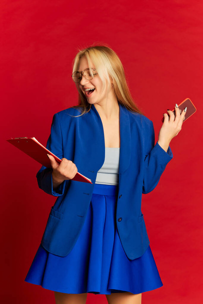 Pozytywna młoda kobieta biznesu lub szczęśliwy student w niebieskim garniturze pozowanie na jasnoczerwonym tle. Startup, motywacja, kariera, emocje, koncepcja osiągnięć. Kolory uzupełniające - Zdjęcie, obraz