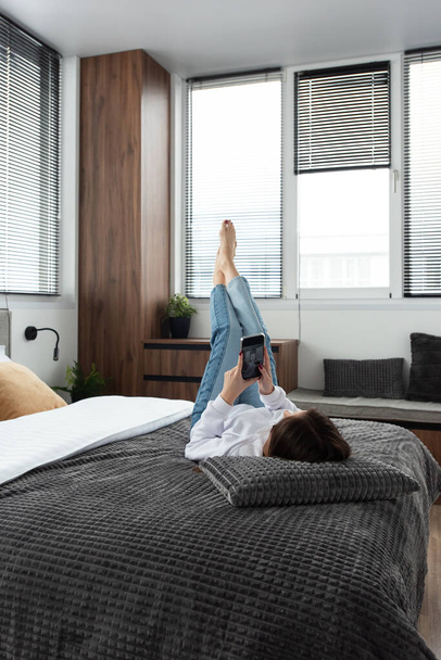 Kobieta z telefonem na łóżku w nowoczesnym mieszkaniu. pokój narożny z żaluzjami na oknach - Zdjęcie, obraz