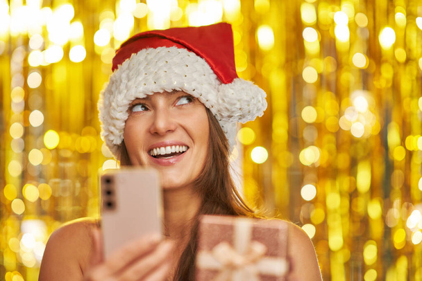 Woman over gold background in Santa Claus hat with phone. High quality photo - Zdjęcie, obraz