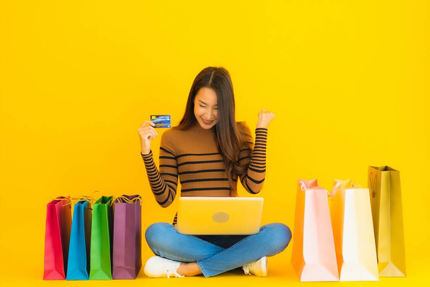 美しい肖像画若いアジアの女性は黄色の背景に買い物袋の周りのオンラインショッピングのためのラップトップやコンピュータ使用クレジットカードで床に座っている - 写真・画像