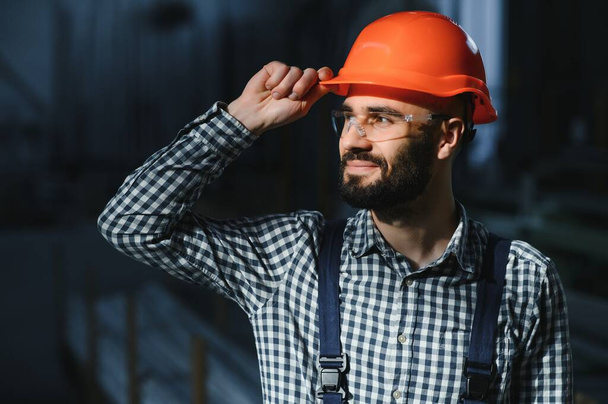 Portrait of Professional Heavy Industry Engineer, Worker Wearing Safety Uniform, Hard Hat Smiling. In the Background Unfocused Large Industrial Factory - Фото, зображення