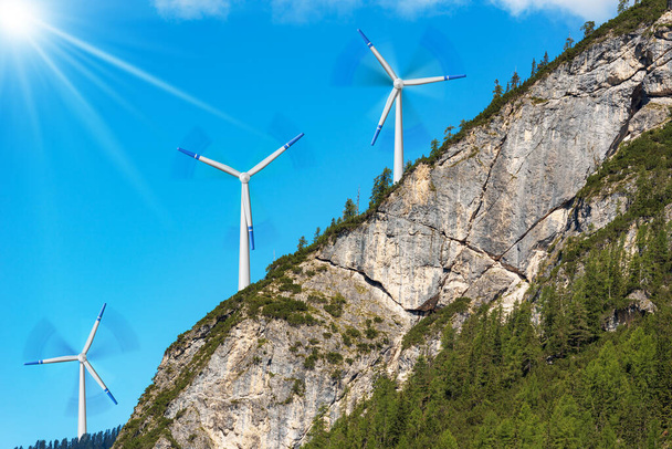 Group of white and blue wind turbines on the top of a mountain rock face, clear blue sky and sunbeams on background. Pine woodland on foreground. Renewable energy concept. - Photo, Image